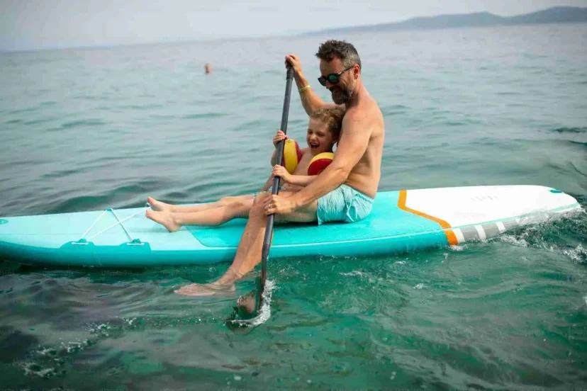 A father and young child, both smiling, sitting on a blue paddleboard in the ocean, with the father paddling."